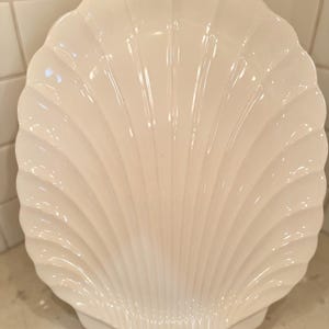 May include: A white, shell-shaped decorative plate. The plate has a scalloped edge and a ribbed design, resembling a seashell. The plate is made of ceramic and is displayed on a countertop. The plate is a decorative item.