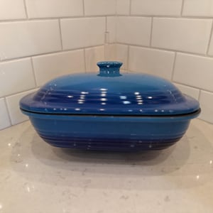 May include: A blue oval-shaped casserole dish with a lid. The dish is a gradient of blues, with a small knob on top. The dish is sitting on a white countertop, with white subway tile in the background.