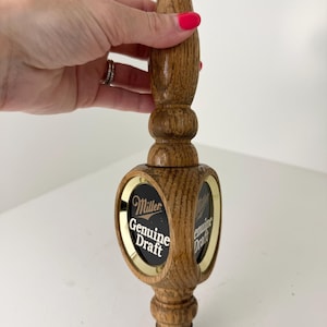May include: A wooden beer tap handle featuring a polished oak finish with a turned, cylindrical design. The center section has an oval, gold-rimmed inlay displaying the black and white Miller Genuine Draft logo and text on both sides.