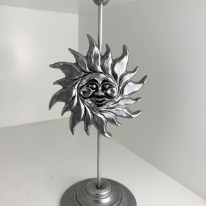 Könnte beinhalten: Ein silberfarbener Kerzenständer aus Metall mit einem zentralen Sonnenmotiv, das ein lächelndes Gesicht und strahlenförmige Flammen zeigt. Der Kerzenhalter steht auf einem runden Fuß mit einem dünnen Stab, der das Ornament und die Tülle trägt.