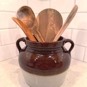 May include: A two-toned ceramic crock with a dark brown glazed upper section and a light cream lower section, featuring two side handles. Three wooden kitchen utensils, including a rounded spoon and a flat paddle, are placed inside the crock.