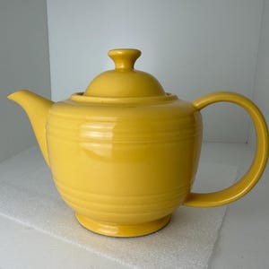 May include: A bright yellow ceramic teapot featuring a rounded body with horizontal embossed ring details, a matching lid with a small knob handle, a curved spout, and a large C-shaped handle extending from the side of the vessel.