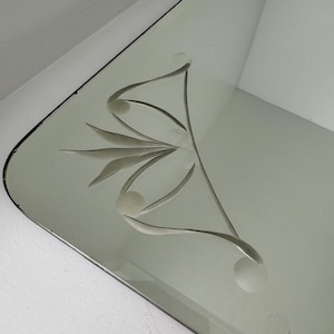 May include: A rectangular wall mirror featuring a beveled edge and a decorative etched floral pattern in the corner. The etching consists of stylized leaves and circular accents, creating a subtle frosted design on the reflective glass surface.