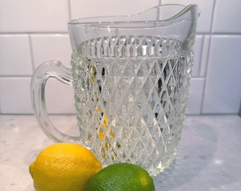 May include: A clear glass pitcher featuring a textured diamond pattern on its exterior, accompanied by one bright yellow lemon and one vibrant green lime placed in front of the vessel. The pitcher has a curved handle and a pouring spout.