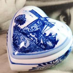 May include: A heart-shaped ceramic trinket box featuring a white base with blue hand-painted illustrations of a traditional windmill, trees, and a house. The lid is decorated with a detailed landscape scene, while the sides feature blue floral patterns.