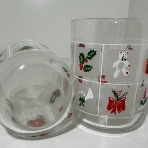 May include: Two clear drinking glasses with Christmas designs. One glass is upright, the other is on its side. The designs include a teddy bear, holly, and a red bow. The glasses are on a white surface.