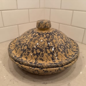 Puede incluir: Una mantequera de cerámica con tapa. El plato es beige con un patrón moteado azul. La tapa tiene un pequeño pomo en la parte superior para facilitar el manejo. El plato está sobre una encimera blanca.