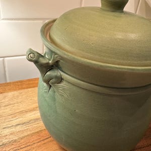 Może przedstawiać: Biały ceramiczny kubek z czarnym uchwytem i napisem "But First, Coffee" pogrubioną, czarną czcionką. Kubek jest ustawiony na rozmytym tle, sugerującym przytulne otoczenie.