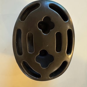 May include: A dark bronze-colored, oval-shaped decorative object with a symmetrical design. The object features a series of elongated openings and four clover-shaped cutouts, creating a pattern of negative space within its form. The surface has a slightly textured appearance.