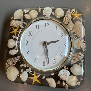 May include: A square wall clock encased in clear resin, featuring a white face with black numerals and two black hands. The border is embedded with various natural seashells and small yellow starfish, creating a textured, multi-dimensional frame.