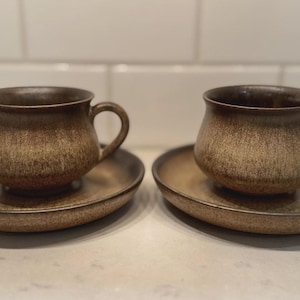 May include: Two matching brown ceramic teacups with saucers. The cups have a rounded shape with a handle, and the saucers are oval. The set has a speckled, textured appearance, with a gradient of brown tones.