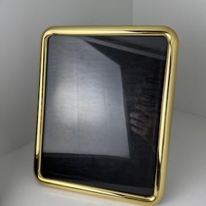 May include: A rectangular picture frame with a polished, reflective gold-tone metal border and rounded corners. The frame features a dark, solid black insert that fills the central area, creating a high-contrast look with the metallic finish.