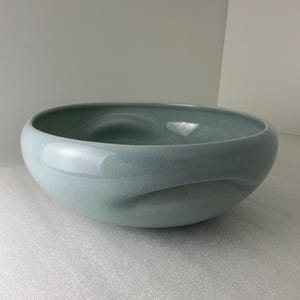 May include: A wide, shallow ceramic bowl with a smooth, glossy finish in a soft, pale blue-green color. The bowl features a distinct, curved indentation along its side, creating a fluid, organic shape with a rounded rim and base.