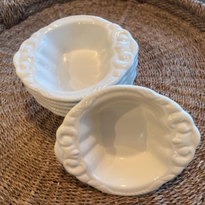 May include: A stack of white, oval-shaped ceramic bowls with a decorative rope-like design around the rim. The bowls are sitting on a woven, brown tray.