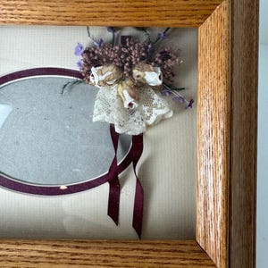May include: A wooden framed oval photo display with a dried flower arrangement. The arrangement includes small white roses, purple accents, and a lace skirt. Burgundy ribbons hang below the arrangement. The frame is made of light brown wood.
