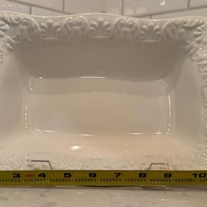 May include: A white rectangular ceramic serving dish with ornate floral detailing around the edges. The dish is approximately 11 inches long. The background features white subway tile and a light-colored countertop.