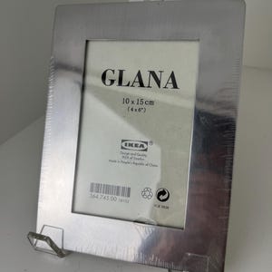 May include: A rectangular silver-colored metal picture frame with a brushed finish. The frame contains a white insert with the text GLANA, 10 x 15 cm (4 x 6 inches), and IKEA branding. The frame is supported by a clear plastic stand.