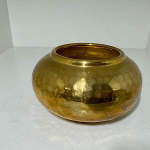 May include: A hammered gold-colored bowl with a wide opening. The bowl has a rounded shape and a textured surface. The interior of the bowl is also gold. This decorative bowl could be used for display or storage.