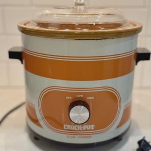 Puede incluir: Una olla de cocción lenta Crock-Pot vintage con tapa de cristal transparente. El electrodoméstico blanco y naranja tiene un dial con ajustes para apagado, bajo y alto. Las palabras "Crock-Pot" y "Slow-Cooker" están impresas en la parte delantera.