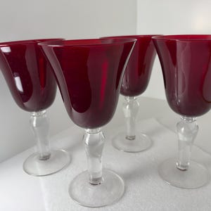 Puede incluir: Un juego de cuatro copas de vino con cuencos de color rojo intenso translúcido y tallos de vidrio transparente con bases circulares. Los cuencos tienen una abertura ancha y acampanada que se estrecha hacia el tallo, creando una silueta elegante.