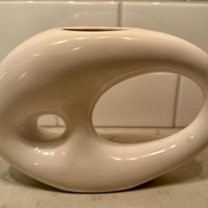 May include: Cream-colored ceramic vase with a unique, organic shape. The vase features a large, oval opening and a smaller, circular opening, creating a distinctive design. The glossy finish enhances the smooth, flowing lines of the decorative piece.