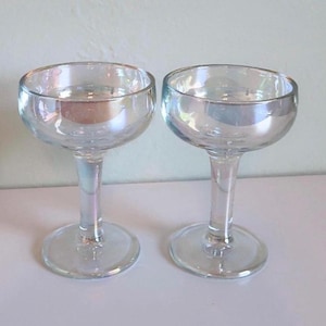May include: A pair of clear, iridescent champagne coupe glasses. Each glass features a rounded bowl, a slender stem, and a circular base. The glasses are placed next to each other, reflecting light and creating a subtle rainbow effect.