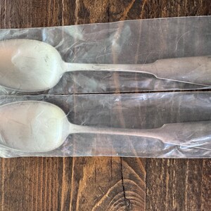 May include: Two silver-toned metal spoons, each individually encased in a clear plastic sleeve. The spoons feature a smooth, oval-shaped bowl and a flat, tapered handle with a rounded, heart-shaped decorative detail at the very end of the stem.