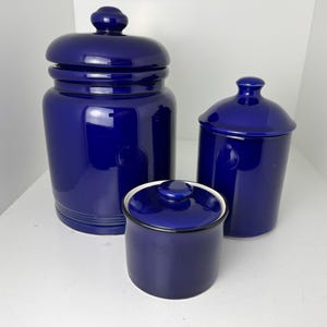 May include: A set of three glossy cobalt blue ceramic canisters with matching lids. The collection includes one large, one medium, and one small cylindrical container, each featuring a smooth finish and a rounded knob handle on the lid.
