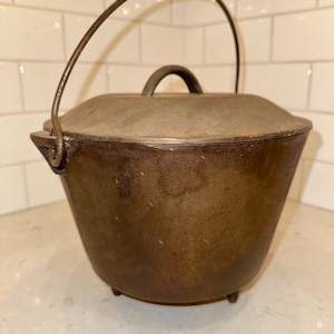 May include: A vintage cast iron Dutch oven with a lid and a curved handle. The pot is dark brown with a slightly textured surface and three small feet. The handle is made of metal.
