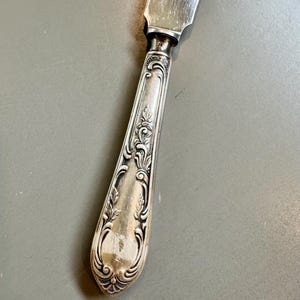 Puede incluir: Un cuchillo de metal plateado con un mango ornamentado y en relieve que presenta intrincados motivos de volutas y hojas. El mango se estrecha hacia una virola lisa y pulida, conectándose a una hoja plana y reflectante con el número 30.