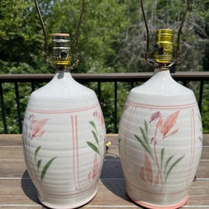 May include: A pair of off-white ceramic table lamps with a ribbed texture, featuring hand-painted pink floral motifs and vertical pink stripes. Each lamp includes a brass-colored metal socket and a harp attachment for a lampshade.