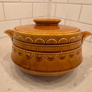 May include: A vintage, lidded ceramic casserole dish in a warm, amber-brown hue. The dish features a scalloped design around the midsection and on the lid, with small handles on either side. The lid has a small, round knob for easy handling.