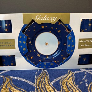 Op de afbeelding: Een boxed set van vakantie servies, met vier kopjes en schotels. De set heeft een marineblauwe achtergrond met gouden sterontwerpen. De doos leest "Galaxy" en "Holiday Dinnerware".