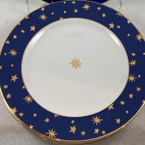 May include: A round dinner plate featuring a white center with a single gold star at the middle. The wide rim is deep blue and decorated with numerous gold stars of varying sizes. The outer and inner edges of the rim are trimmed in gold.