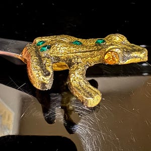 May include: A gold-toned frog figurine with green and orange gemstone accents. The frog has a textured surface and is posed in a crawling position. The figurine is likely a decorative item or a piece of jewelry.