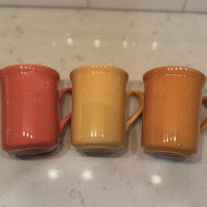 May include: A set of three ceramic mugs with a speckled, matte finish. The mugs are in shades of cream, light gray, and dark gray. Each mug has a simple, modern design and a rounded handle.