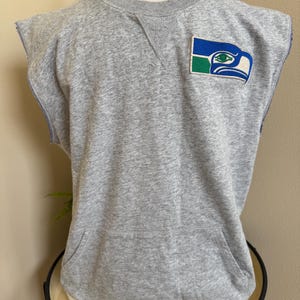 May include: A sleeveless gray sweatshirt with a Seattle Seahawks logo patch. The patch features a blue, green, and white design of a stylized eagle head. The sweatshirt has a front pocket and a crew neck.