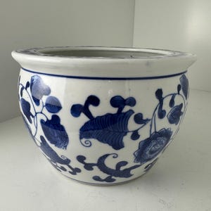 May include: A round ceramic planter featuring a white base with a hand-painted blue floral and vine pattern. The bowl-shaped vessel has a wide, flared rim and a smooth, glossy finish, showcasing intricate botanical motifs encircling the exterior.