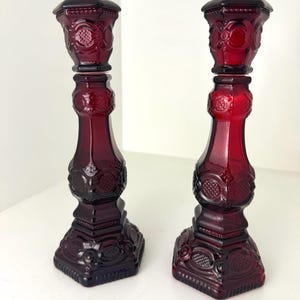 Peut inclure: Une paire de bougeoirs en verre pressé rouge profond présentant un motif géométrique complexe avec des éléments circulaires et hachurés. Chaque bougeoir possède une base hexagonale, une tige effilée et un bord supérieur large.