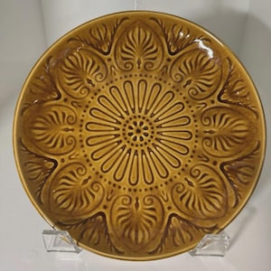 Puede incluir: Un plato redondo de cerámica marrón con un diseño floral en relieve. El plato presenta un patrón central en forma de sol rodeado de hojas y espirales estilizadas. El plato se exhibe sobre un soporte de acrílico transparente.