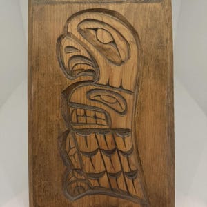 May include: A rectangular, wooden carving featuring a stylized eagle design. The carving is deeply etched into the wood, showcasing the bird's head, beak, and body. The wood has a warm, brown tone, and the carving is likely a piece of Native American art.