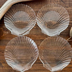 May include: Four clear glass plates shaped like scallop shells. Each plate has a scalloped edge and detailed ridged design. The plates are arranged on a wooden surface, creating a coastal or nautical theme.