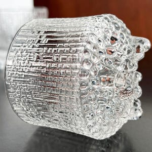 May include: A clear glass tumbler with a textured, bubble-like design. The glass has a cylindrical shape with a slightly flared top. The surface has a rough, uneven texture, creating a unique visual effect. The glass is transparent, allowing light to pass through.