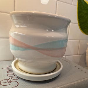 May include: A white ceramic planter with a matching saucer. The planter features a design of pink and blue horizontal stripes. A green and yellow plant is visible in the background. The planter is sitting on a book.