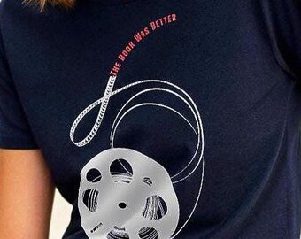 May include: Navy blue t-shirt with a film reel graphic and the text "THE BOOK WAS BETTER" in white and red. The design includes a film reel and a curved line representing film. The shirt has a crew neck.