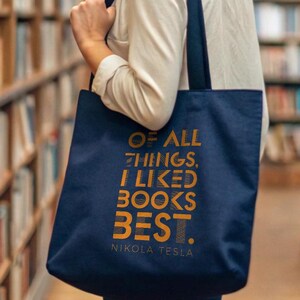 Op de afbeelding: Een marineblauwe draagtas met de quote "OF ALL THINGS, I LIKED BOOKS BEST. - NIKOLA TESLA" in gouden letters. De tas wordt gedragen door een persoon in een bibliotheek, omringd door boekenkasten.