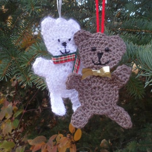 May include: Two crocheted teddy bear ornaments hanging on a Christmas tree. One bear is white with a red and green plaid scarf, the other is brown with a gold bow.