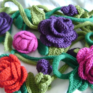 Pode incluir: Uma guirlanda de flores de crochê em tons de roxo, rosa e vermelho, com folhas e caules de crochê verdes.