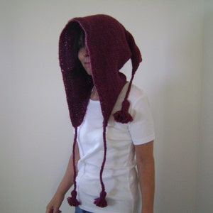 Könnte beinhalten: Ein burgunderfarbener gestrickter Kapuzen-Schal mit langen Bändern und Quasten. Der Schal wird über einem weißen T-Shirt getragen.