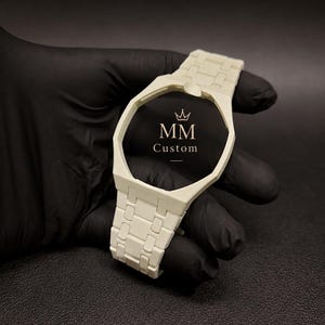 Könnte beinhalten: Ein cremeweißes Uhrengehäuse mit integriertem Gliederarmband und achteckiger Lünette. In der Mitte des Zifferblatts befindet sich ein schwarzer Hintergrund mit einem Kronensymbol über dem goldenen Schriftzug MM Custom, mittig im Rahmen.